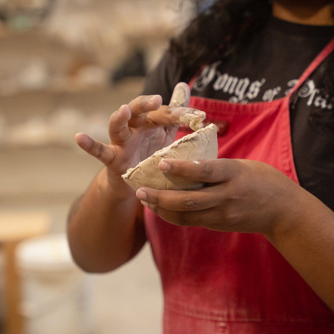 Person in a red apron holding a piece of clay; handbuilding pottery class